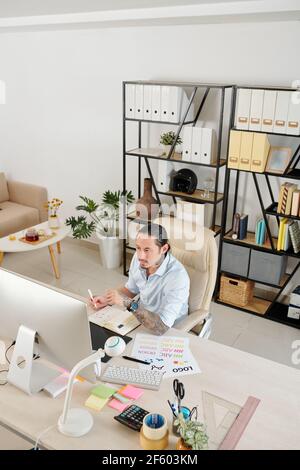 Pensive serious graphic designer working at home office, checking e-mail on computer screen and taking notes in planner Stock Photo