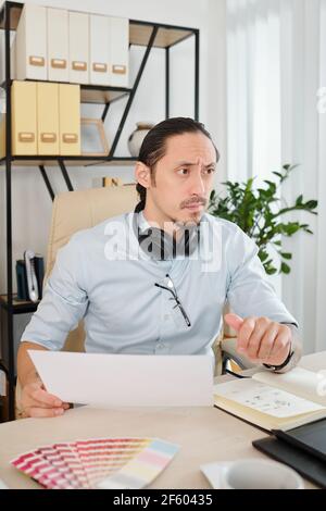 Frowning creative graphic designer working on proejct in home office with opened planner and color palette on desk Stock Photo