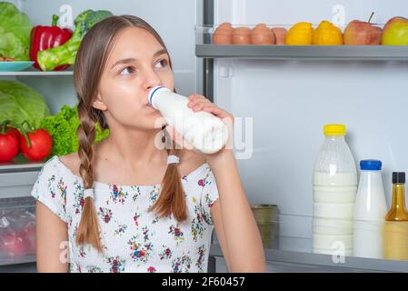 Beautiful young teen girl holding bottle of milk and drinks while standing near open fridge in kitchen at home Stock Photo