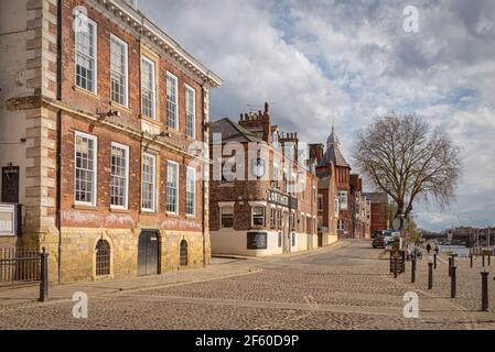 Old red-brick buildings beside the River Ouse in York.  There is a cobbled area in the foreground and a tower on the skyline. Stock Photo