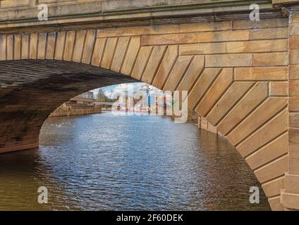An arch of a 19th century stone bridge spanning a river.  Further along the river are pleasure boats and an iron bridge. Birds are on the water. Stock Photo