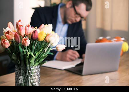 A bouquet of tulips in the foreground. A man wearing a suit and glasses is writing something in a notebook sitting behind a laptop in the background. Stock Photo