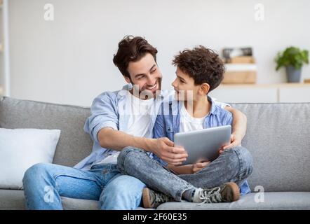 Family time. Happy father teaching his son to use special application on tablet computer, sitting together on sofa Stock Photo
