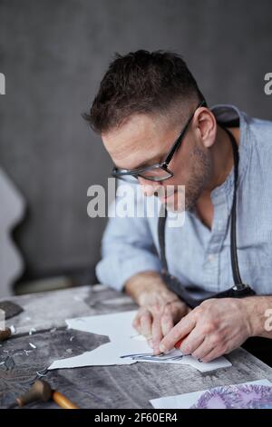Lithograph professional artist handmade worker. Bearded attractive male in apron and eyeglasses making linocut using cutting instrument. High quality photo Stock Photo