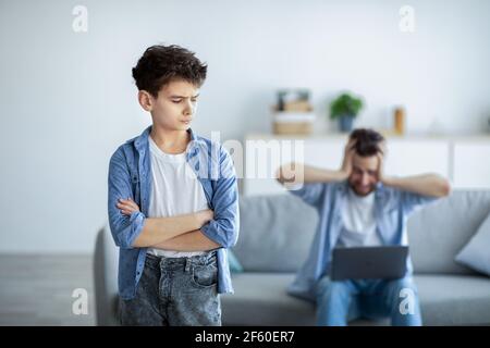 Work at home problem. Boy offended at dad, standing with crossed hands while angry father working on laptop Stock Photo