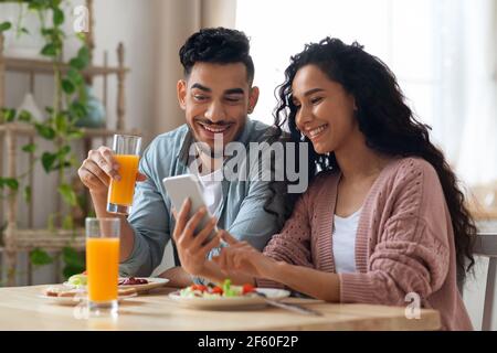 Middle eastern couple watching photos on smartphone while having breakfast in kitchen Stock Photo