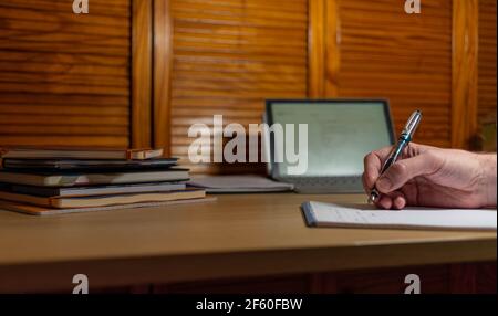 a person taking notes on a pad of paper from a screen. a stack of notebooks to the side Stock Photo