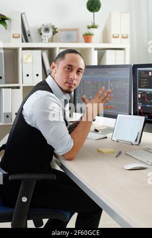 Portrait of serious mixed-race trader with earbuds sitting at his workplace in office and looking at camera Stock Photo