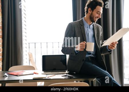 Handsome joyful businessman smiling and examining documents while drinking coffee indoors Stock Photo