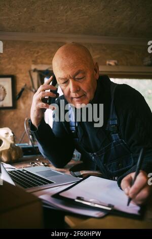 Gay man writing on paper while talking over smart phone at workshop Stock Photo