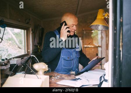 Senior businessman talking on mobile phone while looking at clipboard at garage Stock Photo