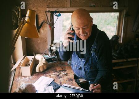 Smiling businessman talking on smart phone in workshop Stock Photo