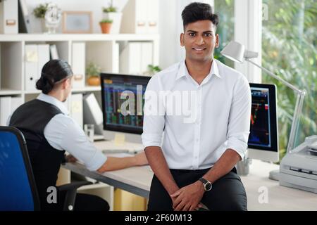 Portrait of happy handsome young Indian stock broker leaning on office table and smiling at camera Stock Photo