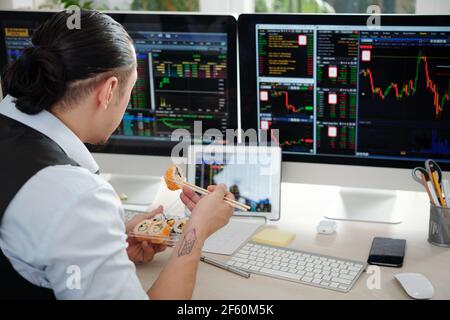 Anxious financial broker eating sushi and analzying stock market trends on computer screens Stock Photo