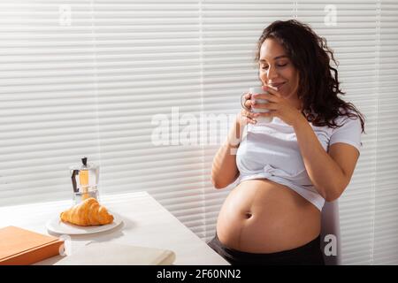 Joyful pregnant young beautiful woman looks through blinds during her morning breakfast with coffee and croissants. Concept of good morning and Stock Photo