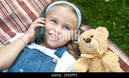 Little happy child girl laying on green lawn with her teddy bear talking on mobile phone. Stock Photo