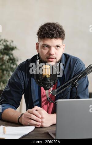 Young male radio dj sitting at the table with laptop and speaking in microphone during his work Stock Photo