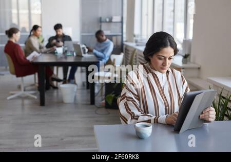 Portrait of contemporary Middle-Eastern businesswoman using digital tablet while working at desk in office with team in background, copy space Stock Photo