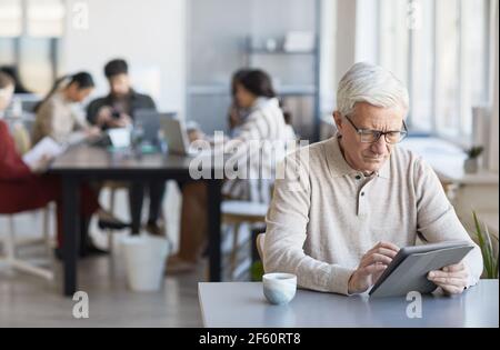 Portrait of contemporary senior man using digital tablet while working at desk in office with team in background, copy space Stock Photo