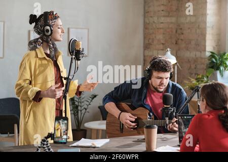 Musical group playing guitar and singing song and performing during air on radio with radio dj sitting opposite them Stock Photo