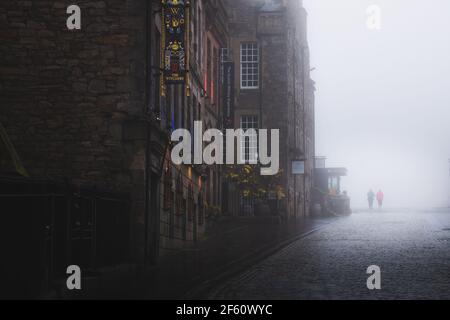 Edinburgh, Scotland - June 16 2020: Two silhouettes emerge in moody atmospheric old town Edinburgh along the cobblestone Royal Mile in misty fog. Stock Photo
