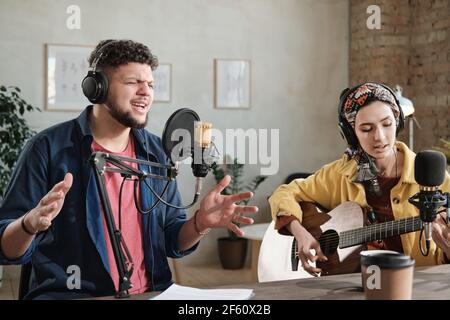 Young singer singing in microphone with young woman playing guitar in recording studio Stock Photo