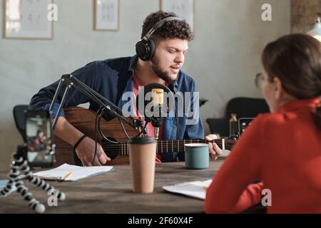 Young man in headphones playing guitar and performing on radio with radio dj sitting opposite him Stock Photo