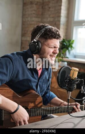 Young man in headphones singing and playing guitar during broadcasting on the radio Stock Photo