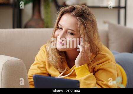 Pretty smiling young blond woman wearing wireless earbuds when watching movie or show online on tablet computer Stock Photo
