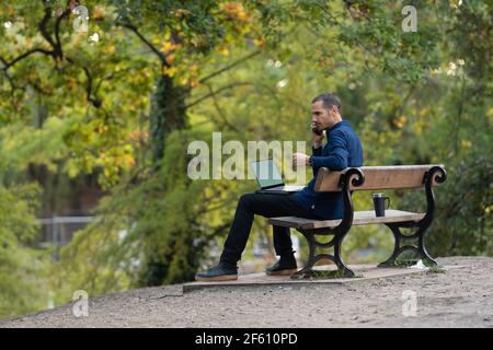Man on park bench, taking call and working on laptop Stock Photo