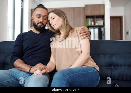 Sad young mixed-race couple reassuring each other when sitting on sofa at home Stock Photo