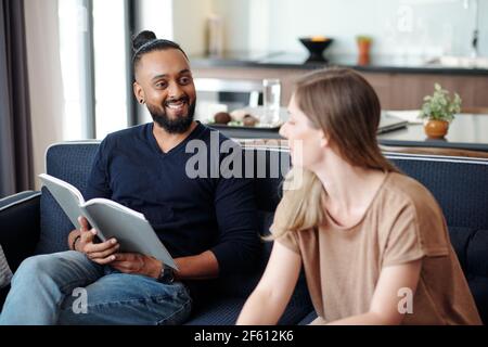 Smiling handsome mixed-race man with open book looking at talking wife Stock Photo
