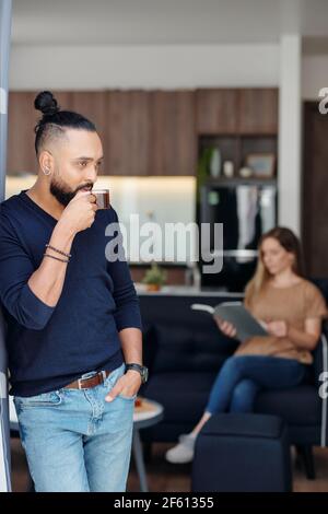 Pensive serious man drinking morning coffee and looking through window, his wife is reading a book in background Stock Photo