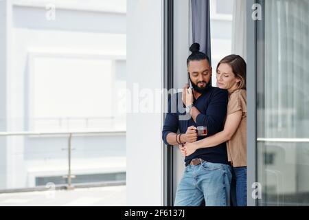 Pretty smiling young woman hugging husband talking on phone with colleague or employee and drinking morning coffee Stock Photo