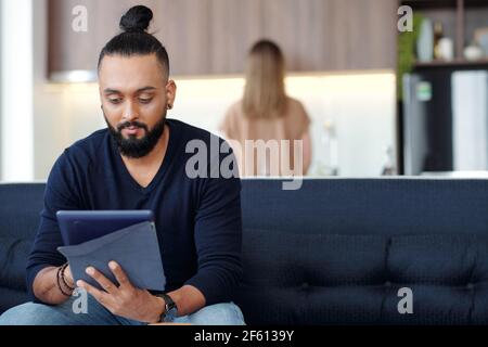 Handsome bearded young man sitting on sofa at home and reading article on tablet computer Stock Photo