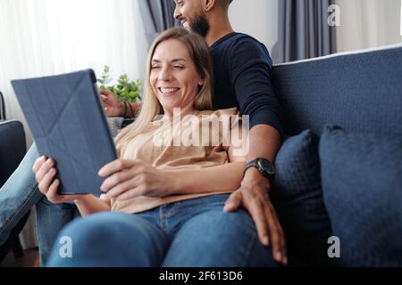 Happy pretty laughing young woman leaning on husband when reading funny stories or watching comedy movie on digital tablet Stock Photo