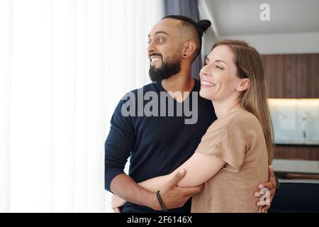 Portrait of happy embracing mixed-race couple hugging and looking through window Stock Photo