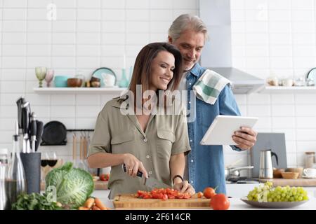 Beautiful couple using a digital tablet and smiling while cooking in kitchen at home. Stock Photo