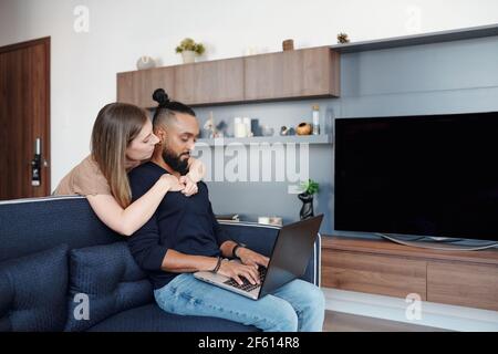 Pretty young woman hugging her boyfriend from behind when he is working on laptop when sitting on sofa at home Stock Photo
