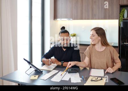 Smiling young woman pointing at bills on table and explaining boyfrend how to manage budget Stock Photo