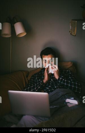 Sick man blowing nose while using laptop in bedroom at home Stock Photo