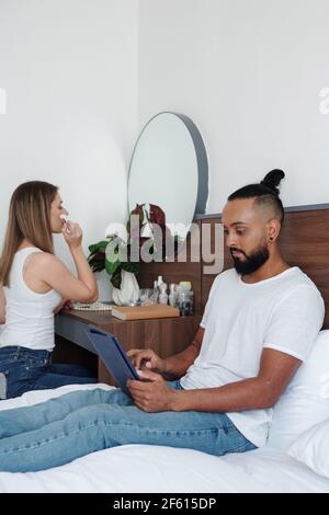 Serious man sitting on bed and reading article on tablet computer when his girlfriend doing make-up at her vanity Stock Photo