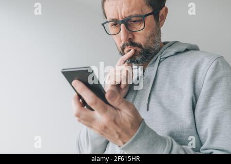 Serious thoughtful mid-adult male looking at mobile smart phone screen with finger on lips and reading text message, close up with selective focus Stock Photo