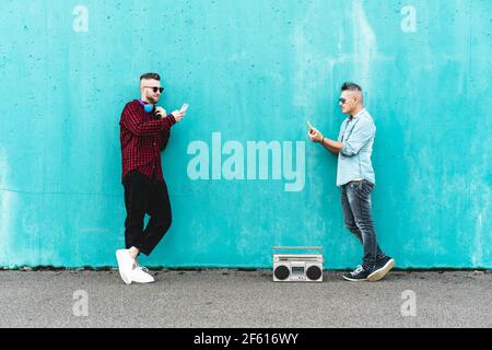 Couple of men using mobile phone against a blue wall background - Concept of falling in love using technology, social networks and mobile phone - LGBT Stock Photo