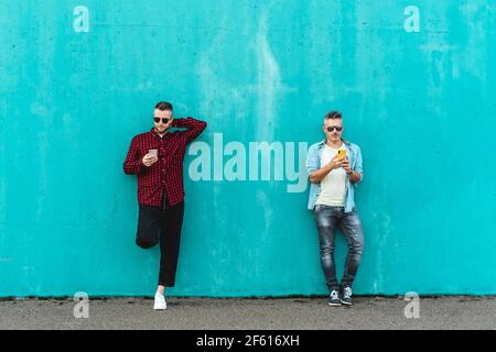 Couple of men using mobile phone against a blue wall background - Concept of falling in love using technology, social networks and mobile phone - LGBT Stock Photo