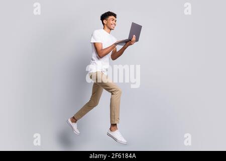 Full size profile photo of brunet optimistic curly guy jump hold laptop wear white t-shirt pants isolated on grey background Stock Photo