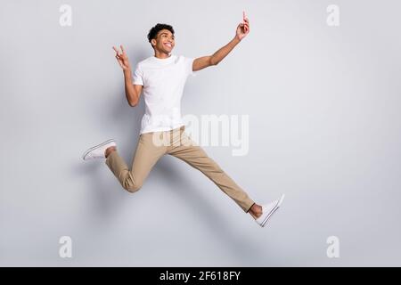 Full size profile photo of brunet optimistic curly guy jump show v-sign do selfie wear white t-shirt pants isolated on grey background Stock Photo