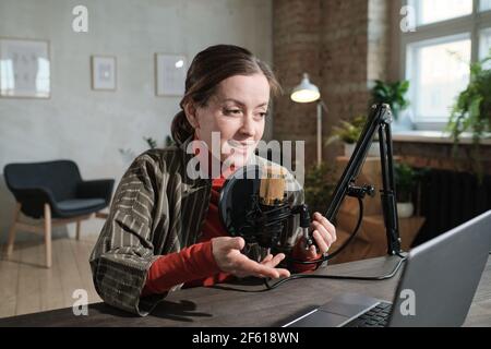 Female radio dj sitting at the table with laptop and using microphone to talk to the listeners in radio studio Stock Photo