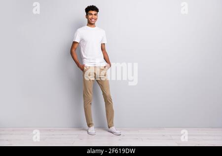Full size photo of brunet optimistic curly guy stand wear white t-shirt trousers sneakers isolated on grey color background Stock Photo
