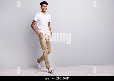 Full size photo of brunet optimistic curly guy go wear white t-shirt trousers sneakers isolated on grey color background Stock Photo